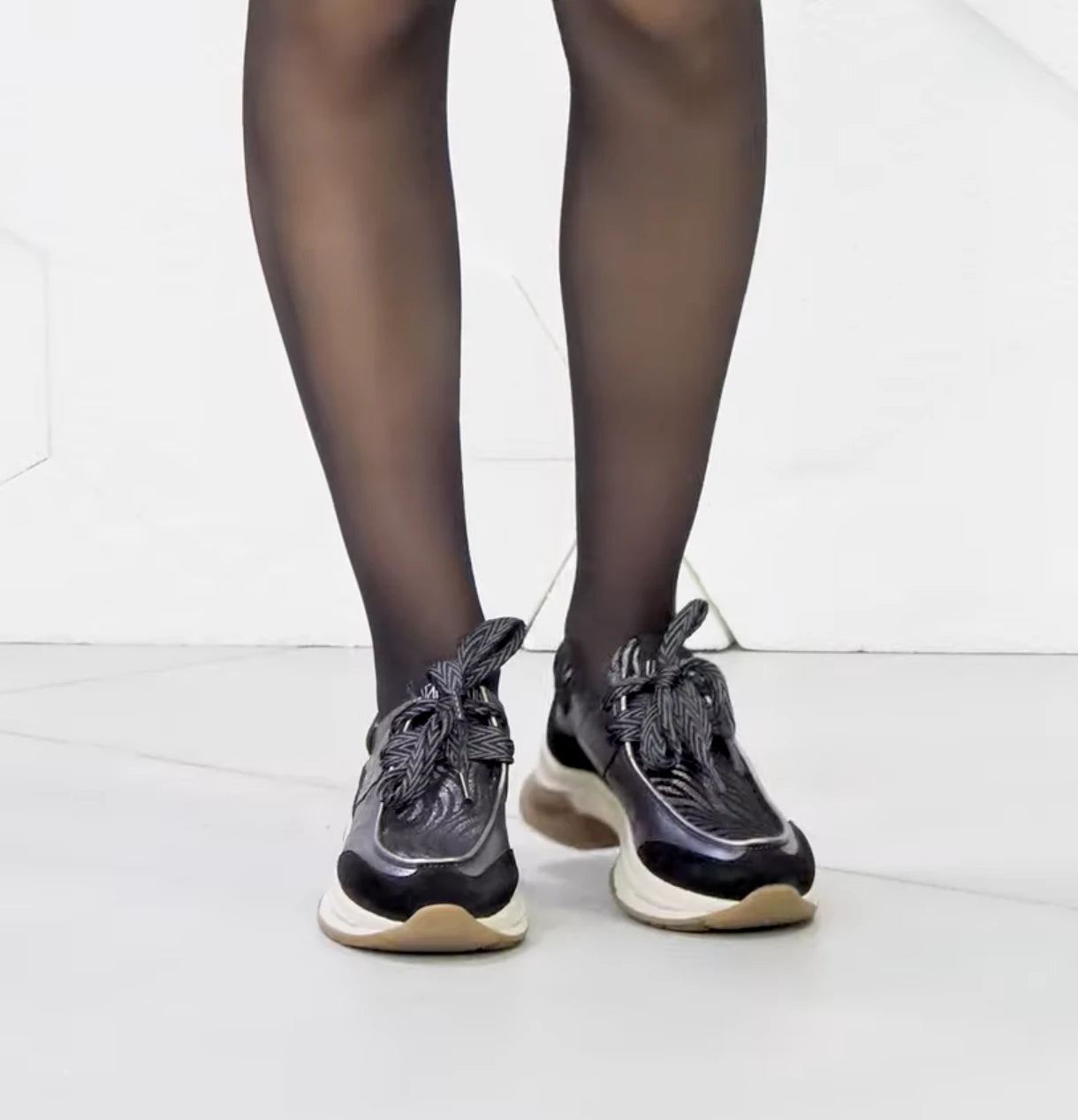Black sneakers with white soles worn with sheer black tights on a white background