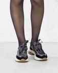 Black sneakers with white soles worn with sheer black tights on a white background