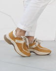 Brown sneakers with white laces worn with white pants on a light gray floor.