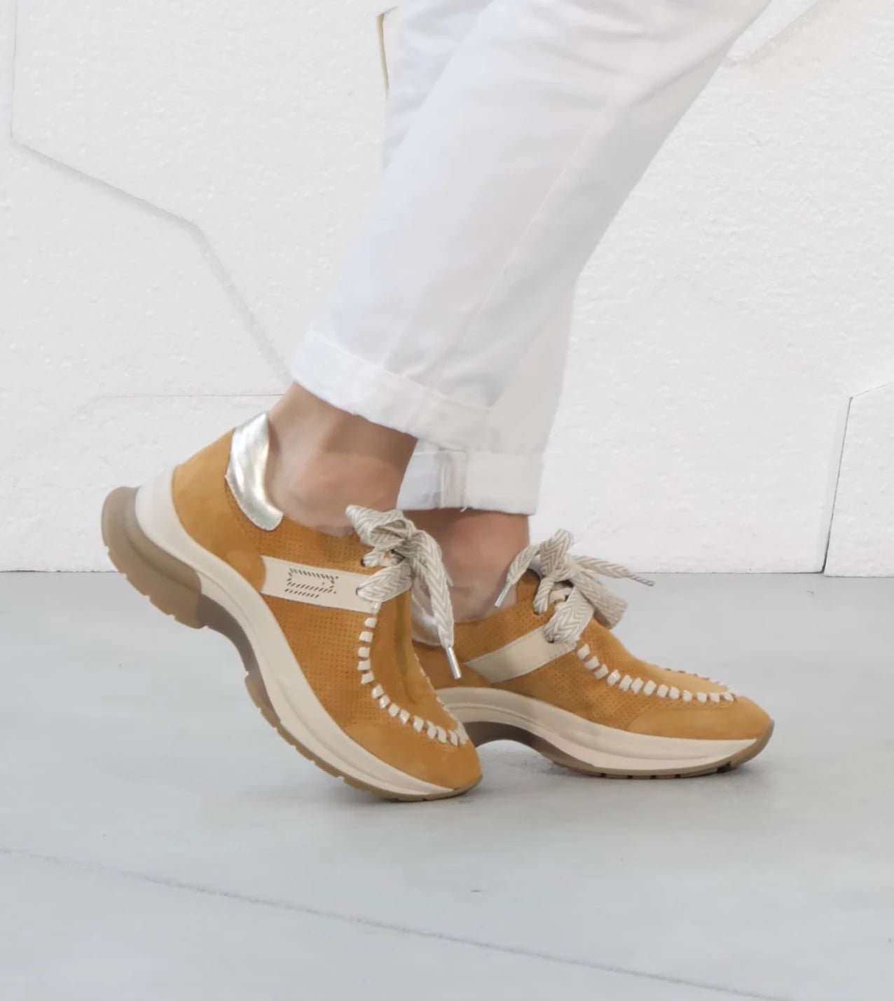 Brown sneakers with white laces worn with white pants on a light gray floor.